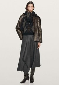 Giacca di pelle marrone, maglione nero con motivo, gonna grigia asimmetrica, stivali neri alti fino al ginocchio, esposti su uno sfondo neutro.