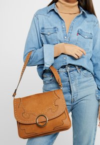 Hnědá kabelka z umělé kůže se silikonovými detaily, kulatým kovovým prvkem a klopou. Obsahuje ramenní popruh a texturovaný povrch.