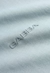 Tissu bleu clair à surface texturée, avec un logo en relief inversé portant l'inscription "GABBA" dans une couleur assortie.