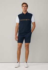 Navyblaues Poloshirt mit weißen Ärmeln, mit goldener "HACKETT"-Markierung. Kombiniert mit navyblauen Shorts und weißen Sneakern. Einfaches, lässiges Design.