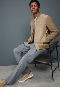 Beige Bomberjacke mit texturierten Ärmeln, graue Hose und beige Sneaker. Modell sitzt auf einem Stuhl vor einem gedämpften grauen Hintergrund.