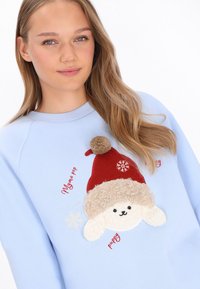 Világoskék pulóver puha fehér kutyát ábrázol piros sapkával, hímzett részletekkel és bojtos díszítéssel. Hópelyhek és felirat teszik teljessé a mintát.