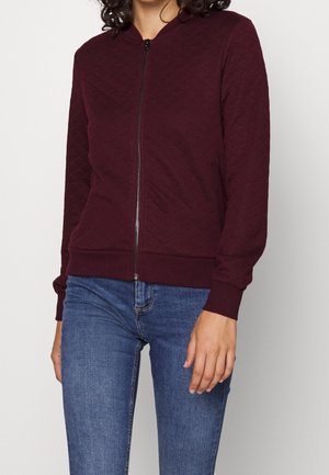 Femme portant une veste matelassée bordeaux avec fermeture éclair, manches et ourlet côtelés, associée à un jean en denim bleu, debout devant un fond neutre.