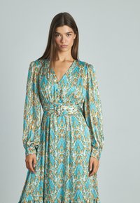Robe portefeuille à manches longues en turquoise avec des motifs orange et crème, dotée d'une taille ceinturée, d'un décolleté en V et d'un ourlet à volants.