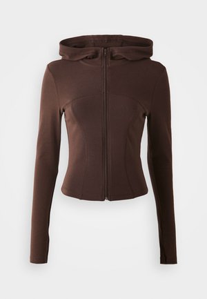 Rudas zip-up džemperis su prigludusia forma, ilgomis rankovėmis ir kapišonu. Pagamintas iš lygaus, tamprios medžiagos, su lenktomis siūlėmis ir sraigtų formą.