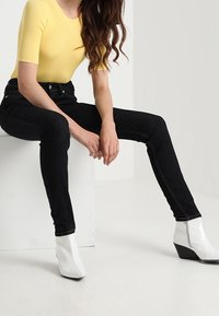 Jeans negros de cintura alta con un corte ajustado, combinados con una blusa amarilla de canalé. Botines blancos con tacón y detalle de cremallera lateral.