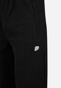 Pantalons noirs texturés arborant un motif à côtes verticales et un petit logo blanc. Le tissu semble doux et est agrémenté de deux poches latérales.