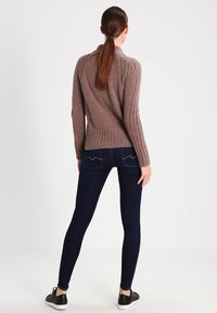 Suéter castanho com riscas, gola alta, corpo ajustado e textura estampada, combinado com jeans escuros e sapatilhas pretas.