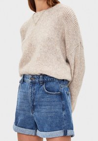 Mujer vistiendo un suéter de punto beige metido en pantalones cortos de mezclilla azul remangados, con un collar de plata visible sobre un fondo liso.