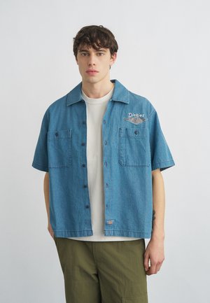 Noor mees lühikeste tumedate juustega, seljas sinine denimmantel, mida ta kannab avatud olekus üle valge t-särgi ja oliivrohelised püksid.