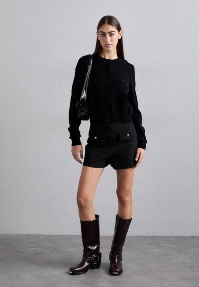 Cardigan nero con scollo rotondo e chiusura a bottoni, abbinato a pantaloni corti neri a vita alta con dettagli dorati. Stivali marroni al ginocchio.