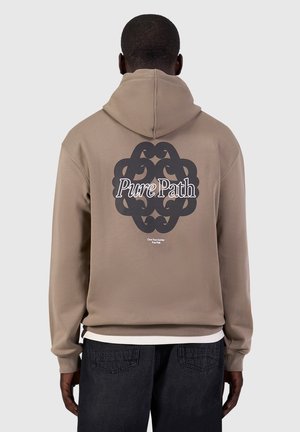 Persoon met de rug naar de kijker, draagt een beige hoodie met groot zwart sierlijk ontwerp en tekst "Pure Path" op de rug, in combinatie met donkere spijkerbroek.