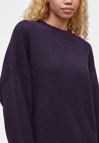 Jeune personne aux cheveux blonds bouclés portant un pull en laine violet foncé ample avec un col rond, debout devant un fond uni.