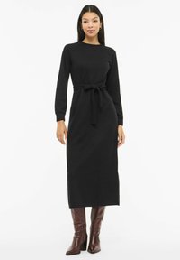 Žena v dlouhých černých šatech s páskem a dlouhými rukávy, stojící v hnědých kozačkách s podpatkem sahajících ke kolenům, na bílém pozadí.