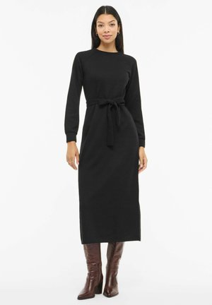 Nainen pukeutuneena pitkään mustaan vyöllä varustettuun mekkoon, jossa on pitkät hihat, ja ruskeat polven korkeudelle yltävät korkokengät, seisomassa valkoista taustaa vasten.