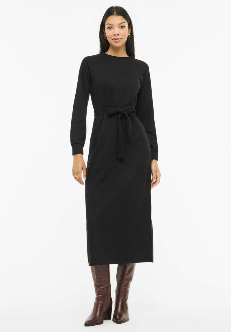 Žena v dlouhých černých šatech s páskem a dlouhými rukávy, stojící v hnědých kozačkách s podpatkem sahajících ke kolenům, na bílém pozadí.