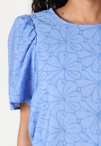 Blouse bleu clair avec des manches bouffantes, une broderie florale complexe, une texture lisse et un col rond.