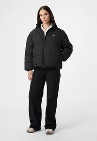 Czarna pikowana kurtka puchowa z przeszyciami, przednim zamkiem błyskawicznym i logo FILA na piersi. Noszona na białej koszuli w połączeniu z czarnymi spodniami.