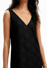 Robe sans manches noire avec une texture en dentelle florale et un décolleté en V profond, accompagnée d'un collier en forme de cœur argenté.