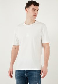 Hvid bomulds t-shirt med korte ærmer og rund halsudskæring, prydet med et diskret præg logo på brystet. Kombineret med blå denim jeans.