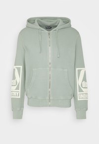 Ljusgrön zip-up hoodie med textilstruktur, två framfickor och vita grafiska tryck på båda ärmarna. Har en huva med dragsko.