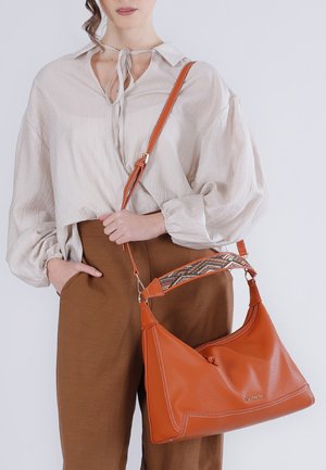 Donna che indossa una camicetta beige larga e pantaloni marroni, che tiene in mano una grande borsa a tracolla arancione in pelle con tracolla decorata.