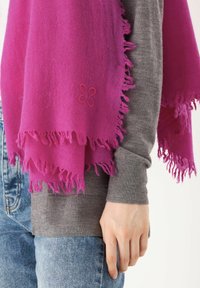 Écharpe fuchsia avec des bords effilochés, fabriquée à partir de matériau doux. Présente un détail de fleur brodé. Portée sur un pull gris avec un jean bleu.
