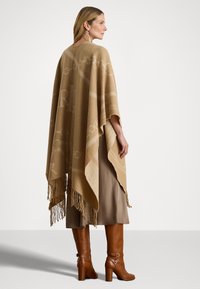 Poncho beige con frange, caratterizzato da motivi tonali. Indossato sopra una gonna marrone e abbinato a stivali alti fino al ginocchio marroni con tacco.