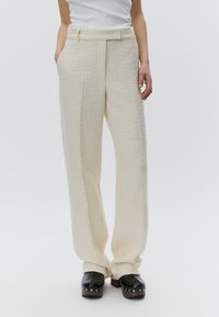 Femme portant un pantalon large texturé cr ème, un haut blanc rentré et des sabots plateformes noirs avec des clous argentés sur les semelles.