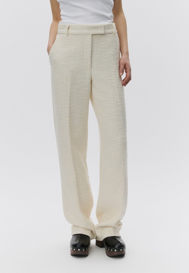 Femme portant un pantalon large texturé crème, un haut blanc rentré et des sabots plateformes noirs avec des clous argentés sur les semelles.