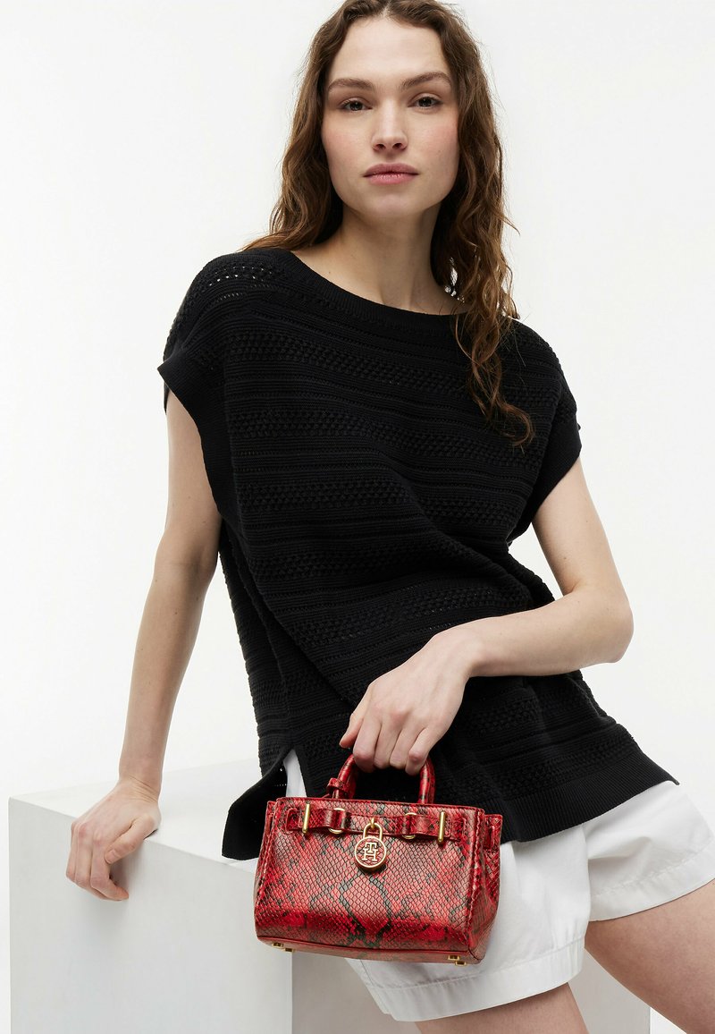 Femme portant un haut en maille noire texturée et un short blanc, assise avec un sac à main en peau de serpent rouge reposant sur sa cuisse.