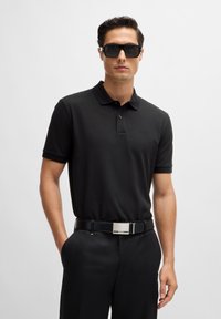 Black polo shirt with a classic collar, short sleeves, and subtle logo. Worn with black trousers and a black belt featuring a silver buckle.