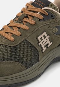 Sneaker verde oliva caratterizzato da materiali in suede e mesh, lacci marroni e un dettaglio del logo con accento beige sul lato.