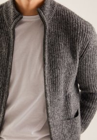 En person som bär en vit T-shirt med rund hals under en delvis zipad tjock grå stickad cardigan med en framficka.