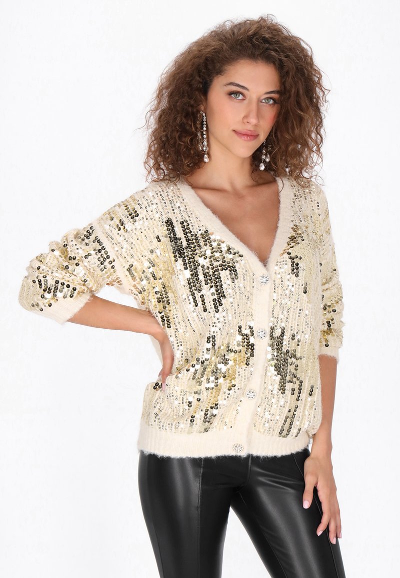 Crème cardi met pailletten in gouden en zwarte patronen, V-hals, knoopsluiting en zachte textuur; gecombineerd met zwarte broek.