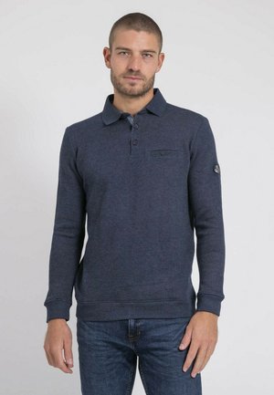 Homme portant un polo à manches longues bleu marine avec col à boutons et un jean bleu, debout devant un fond clair uni.