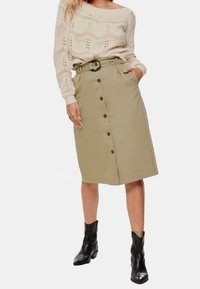 Jupe crayon kaki avec une taille ceinturée, dotée d'une patte de boutonnage à l'avant et de poches latérales. Associée à un pull texturé crème et des bottines noires.