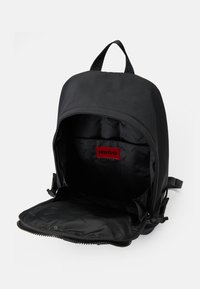 Sac à dos noir avec compartiment principal ouvert révélant une poche intérieure et une étiquette de marque "HUGO" rouge sur le tissu noir.