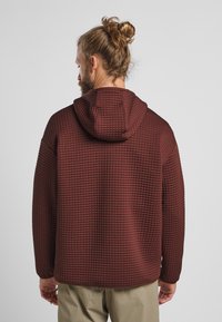 Bordeauxfarbener Kapuzenpullover mit einem strukturierten, gitterartigen Muster. Der Stoff ist dick, mit einer lässigen Passform und überschnittenen Schultern, von hinten gezeigt.