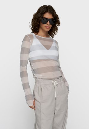 Vrouw met krullend haar die grote zwarte zonnebrillen draagt, een doorschijnende gestreepte top met lange mouwen en lichtbeige broek met trekkoord, handen in de zakken.