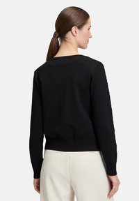 Pull en tricot noir avec un col rond et un ourlet côtelé. Présente des accentuations de boutons le long des manches. Porté avec un pantalon beige.