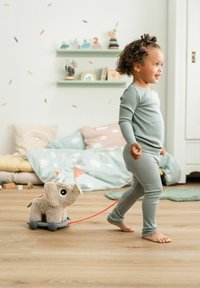 Tout-petit en pyjama vert tirant un jouet éléphant en peluche sur roues à travers un sol en bois dans une chambre décorée.