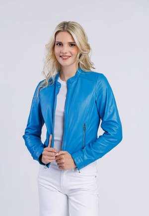 Veste en cuir bleu avec col montant, fermeture éclair devant et poches latérales. Texture lisse et design ajusté, assortie à un haut blanc et un pantalon.