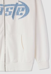 Felpa bianca con zip, tasche frontali e logo con texture in denim blu cucito sul petto, mostrata dalla vita fino a metà manica.