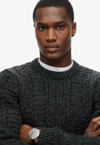 Charcoal grijze gebreide trui met een gestructureerd, geribbeld ontwerp. Het model draagt een witte crewneck shirt eronder en een zwarte horloge om de pols.