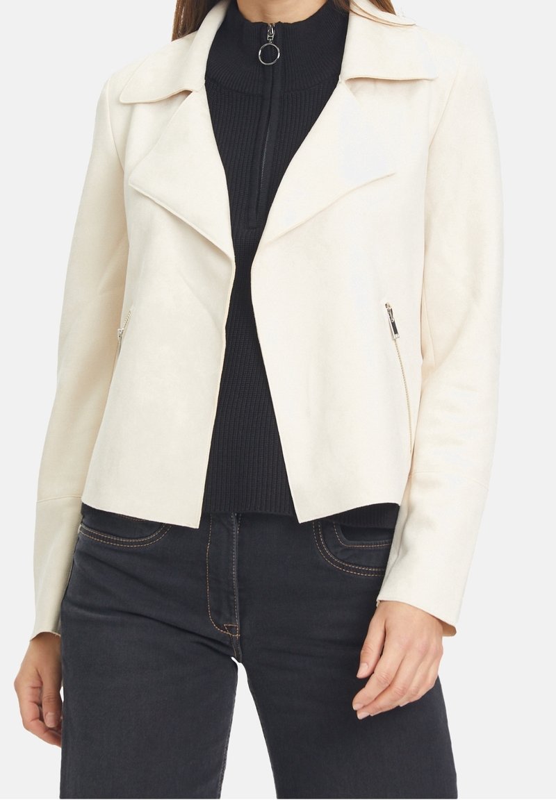 Light cream faux suede jacket with a notched collar, zippered side pockets, and a fitted design, paired with a ribbed black turtleneck.