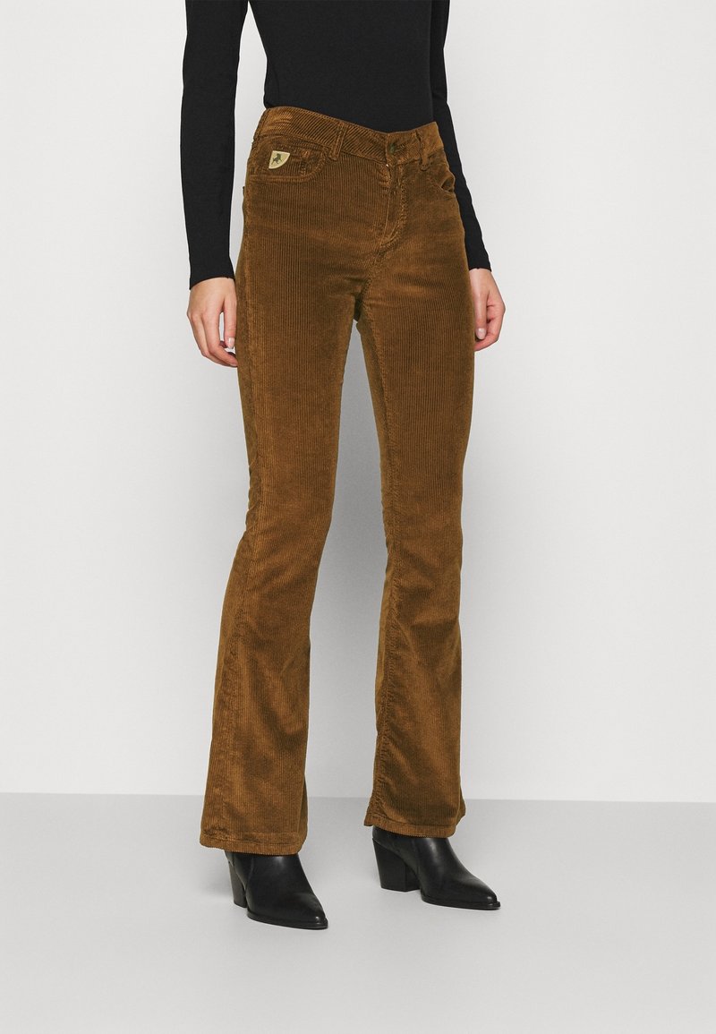 Femme portant un pantalon flare en velours côtelé marron, des bottines noires à bout pointu, et un top noir cintré à manches longues, debout devant un fond uni.