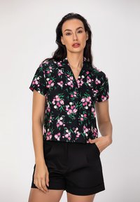 Blouse noire à manches courtes avec un imprimé floral en rose et blanc, dotée d'un col et d'une fermeture à boutons. Associée à un short noir.