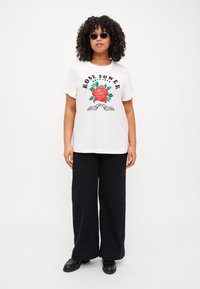 Weißes Baumwoll-T-Shirt mit einem roten Rosenmotiv und grünen Blättern, schwarzer Schrift "ROSE POWER TRUE LOVE", kombiniert mit schwarzen weiten Hosen.