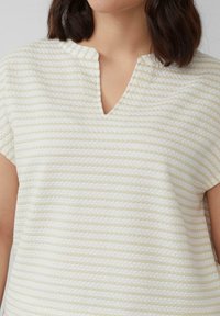 Kobieta w krótkim rękawie, nosząca kremowo-beżowy, teksturowany top w paski z dekoltem w kształcie litery V, z widocznymi włosami sięgającymi ramion w ciemnym kolorze.